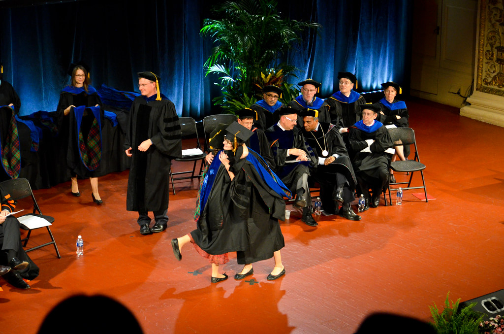A graduate in doctoral robes is hooded on stage while faculty in academic dress sit behind.