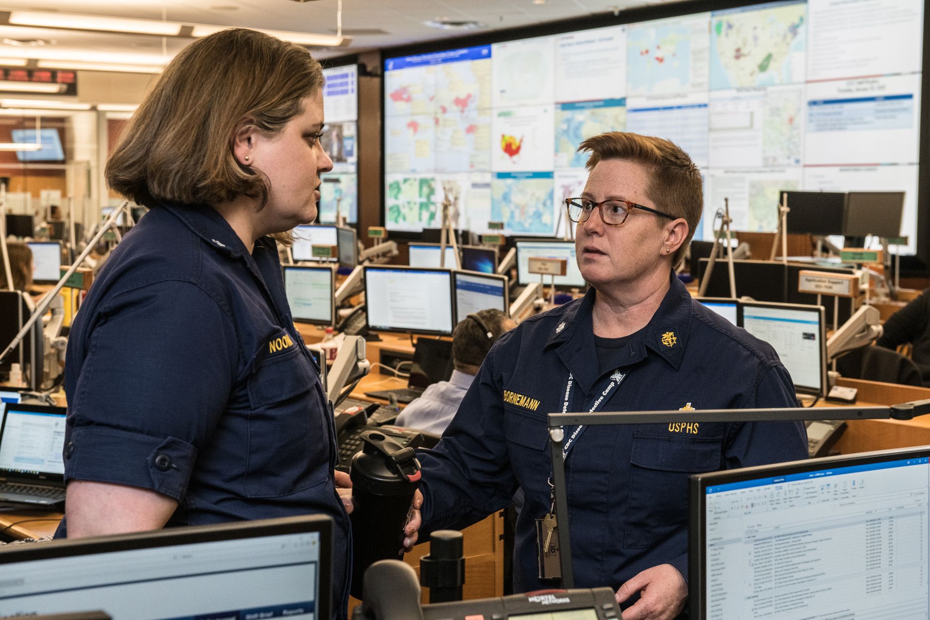 Emergency management staff in uniform working at computer stations in front of wall-mounted maps during an operations center activation